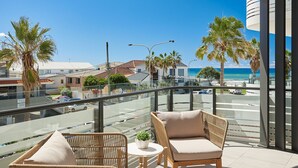 Ein Balkon mit Korbstühlen und einem kleinen Tisch, der auf eine Straße mit Palmen und einen Strand blickt.