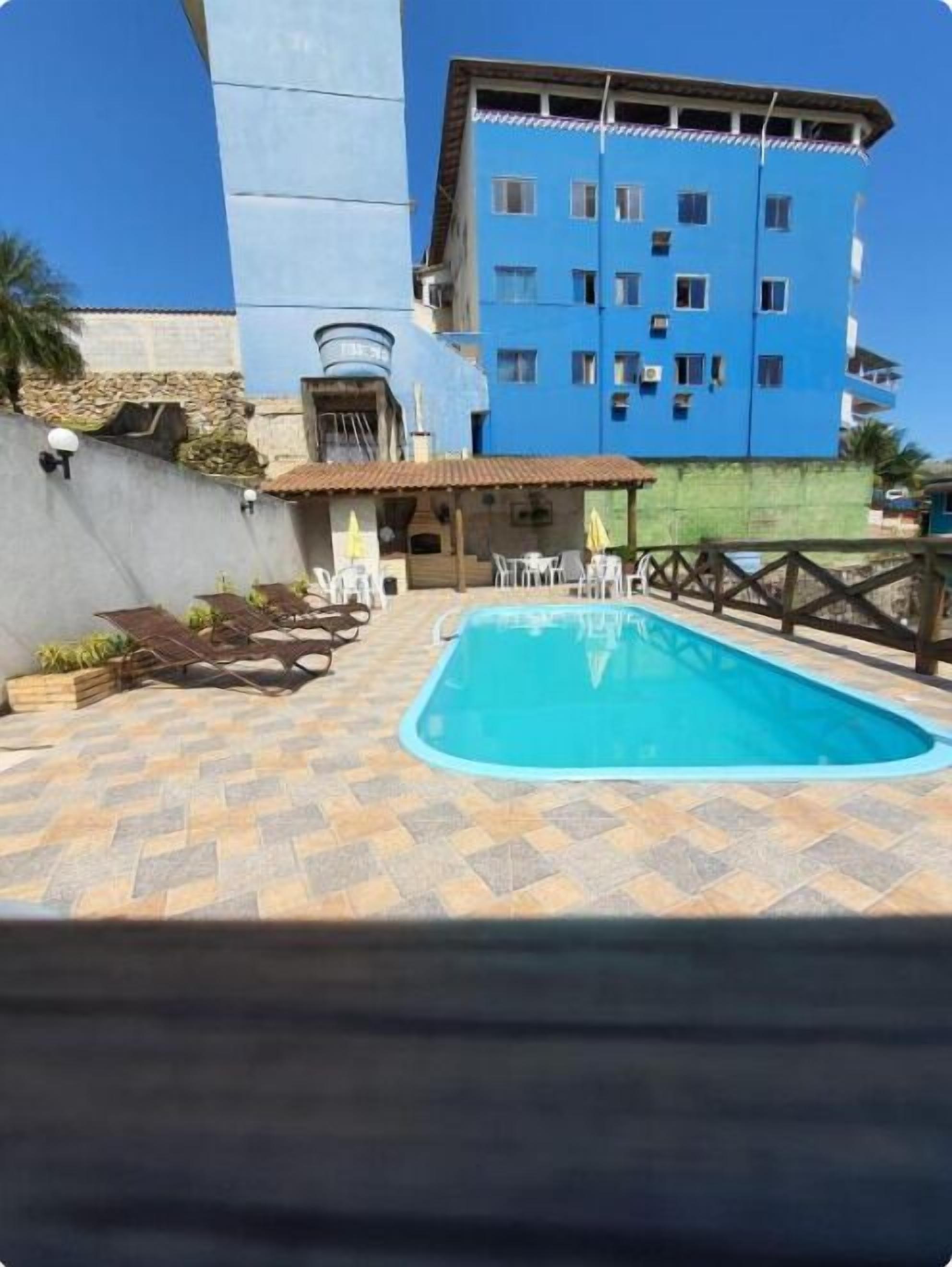 Piscina con suelo de baldosas, un edificio azul al fondo y mobiliario de exterior.