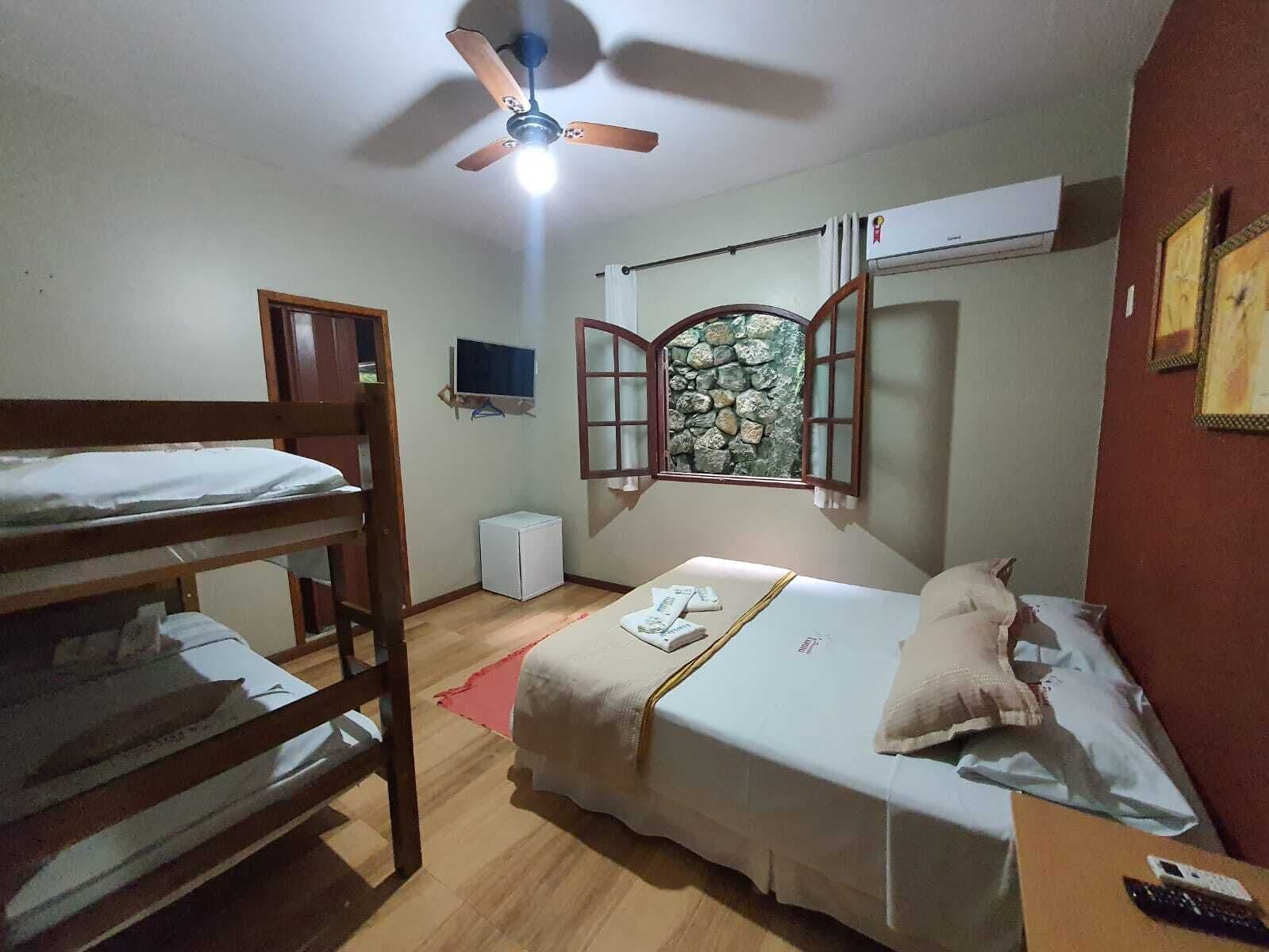 A dormitory room with bunk beds, a ceiling fan, a small TV, a stone wall, and a window with wooden shutters.