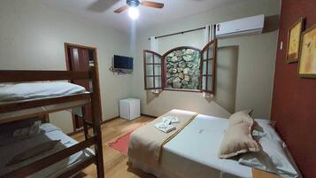 A dormitory room with bunk beds, a ceiling fan, a small TV, a stone wall, and a window with wooden shutters.