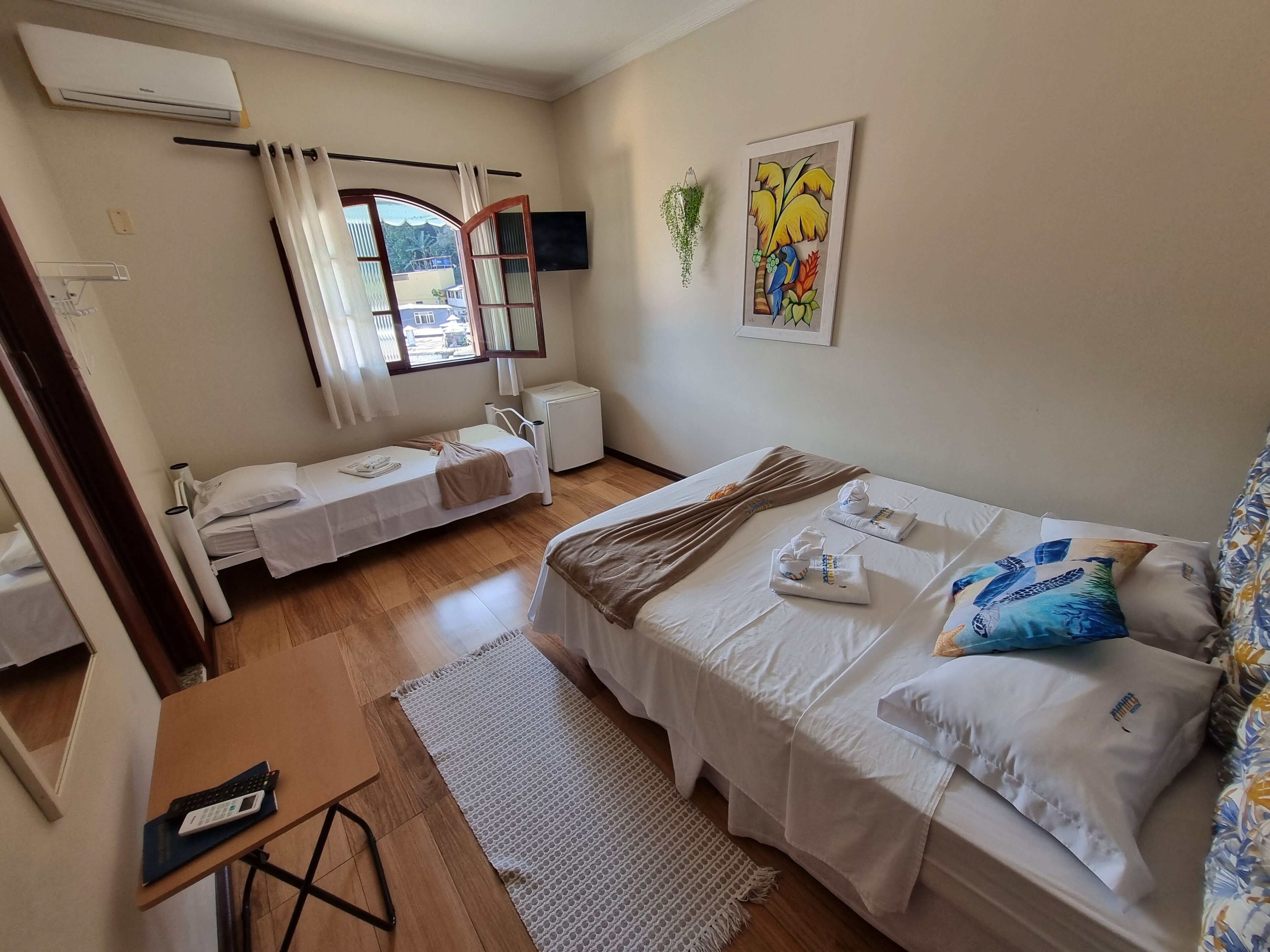 A hotel room with two single beds, a desk with a chair, a small table, a window with curtains, a wall-mounted TV, and a painting.