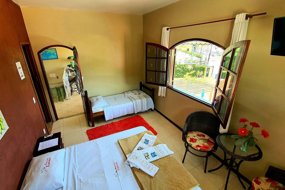 A hotel room with two beds, a small table with a vase of flowers, a chair, and a window with a view of the outside.