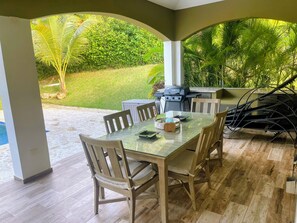 An outdoor patio with a glass-top table, wooden chairs, and a grill.