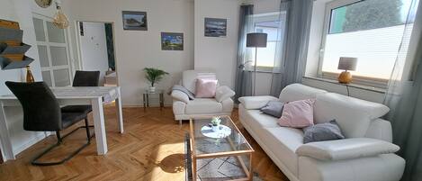 Una sala de estar moderna con un sofá blanco, una mesa de centro de vidrio y un escritorio con una computadora portátil. Hay cuadros en las paredes y una planta en una mesita auxiliar.