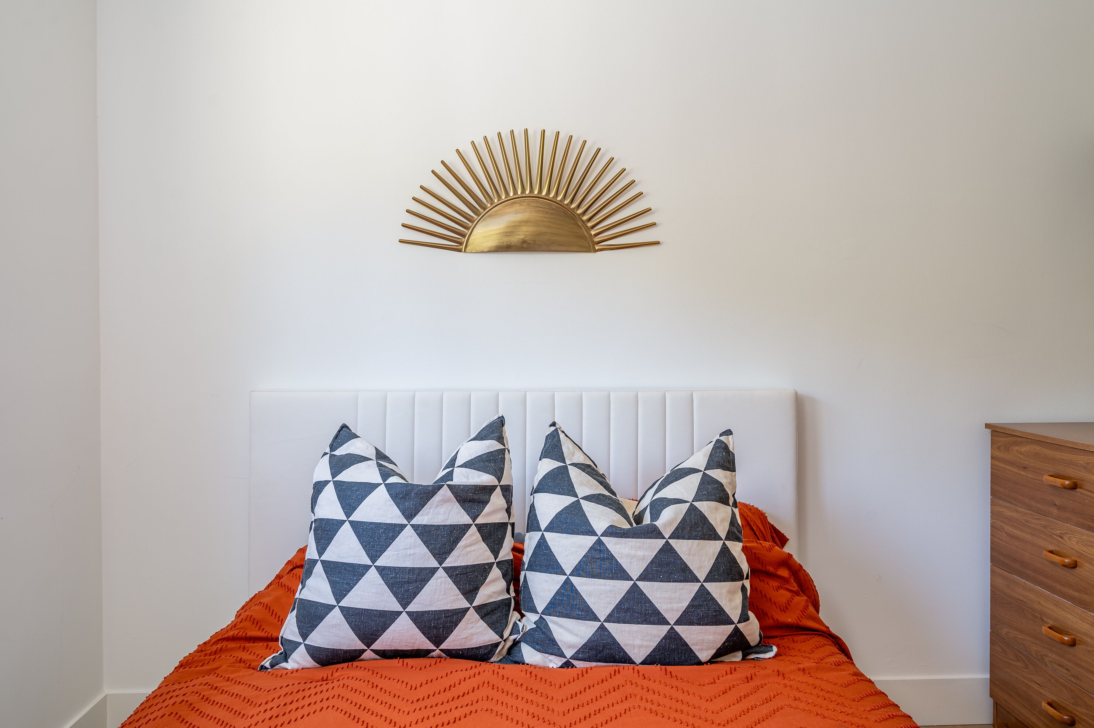 Ein Bett mit weißem Kopfteil, zwei gestreifte Kissen, einer orangefarbenen Decke und einer goldfarbenen Wanddekoration in Form einer Sonne.