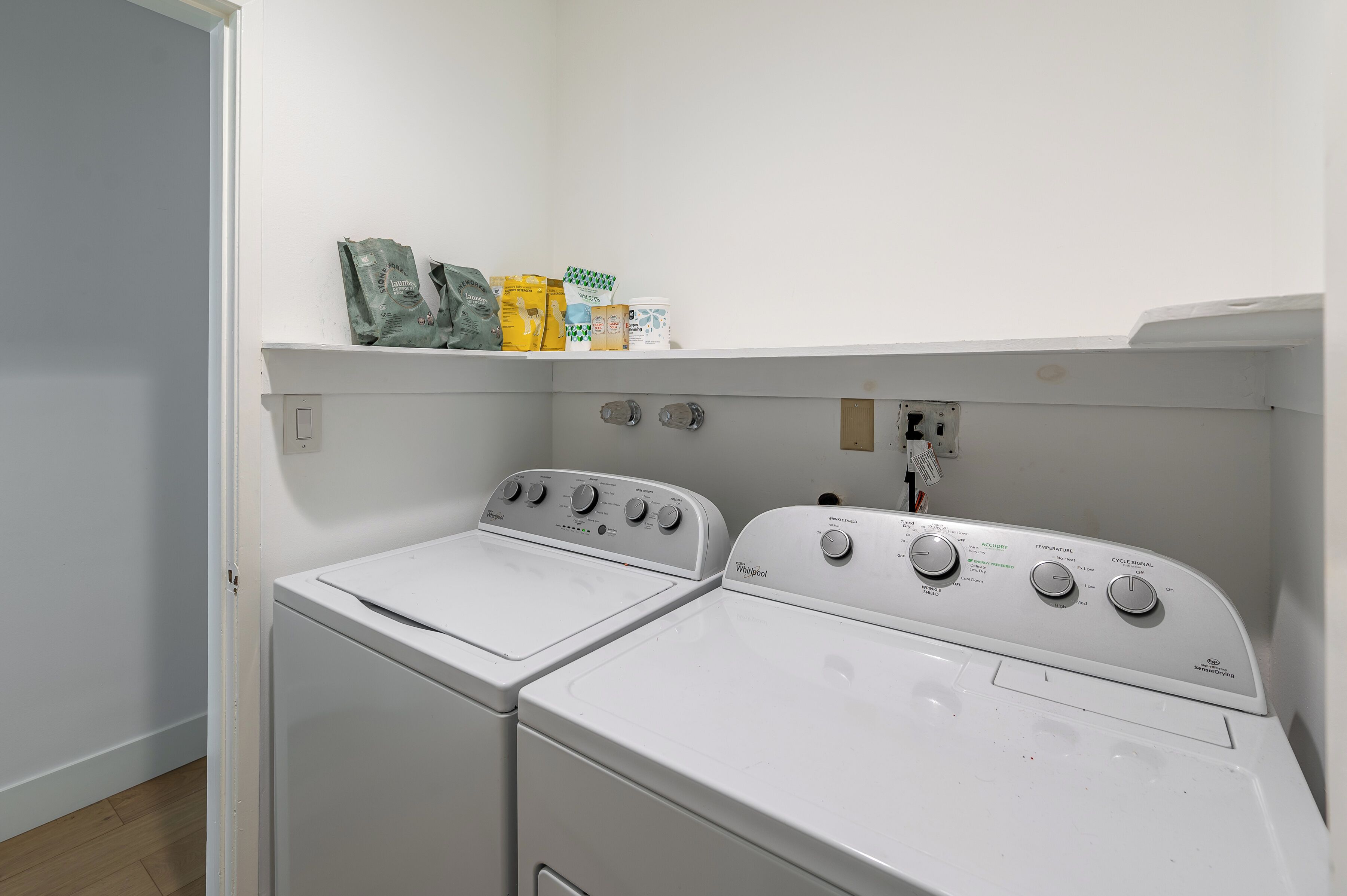 A laundry area with two white Whirlpool washers and a shelf above containing detergent bags.