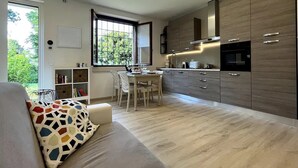 A modern kitchen with wooden cabinets, a dining area with a table and chairs, and a sofa with a colorful cushion. - Cozy and bright apartment with a garden: pets allowed (Lombardia)