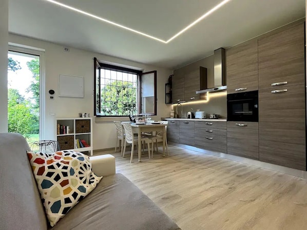 A modern kitchen with wooden cabinets, a dining area with a table and chairs, and a sofa with a colorful cushion.