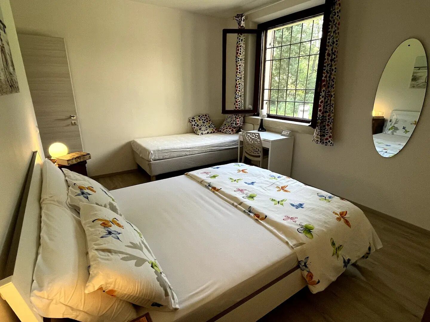 A bedroom with a bed, a desk, a chair, a window with curtains, a mirror, and a small bedside table with a lamp.
