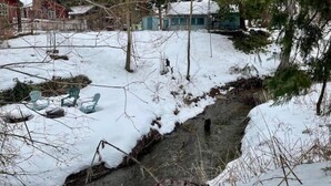 Une cabane avec une cour enneigée, un petit ruisseau et quelques chaises.