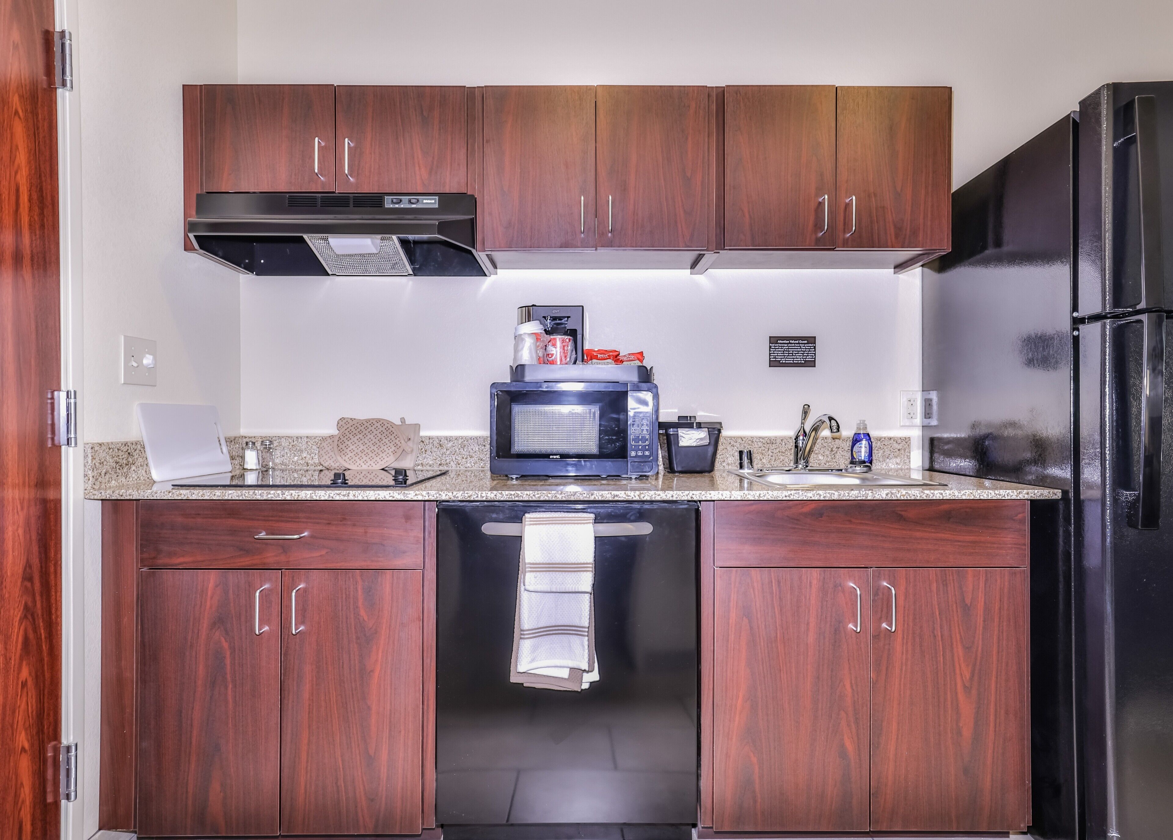 Una cocina compacta con muebles de madera oscura, una campana extractora de acero inoxidable, un microondas y un refrigerador negro.