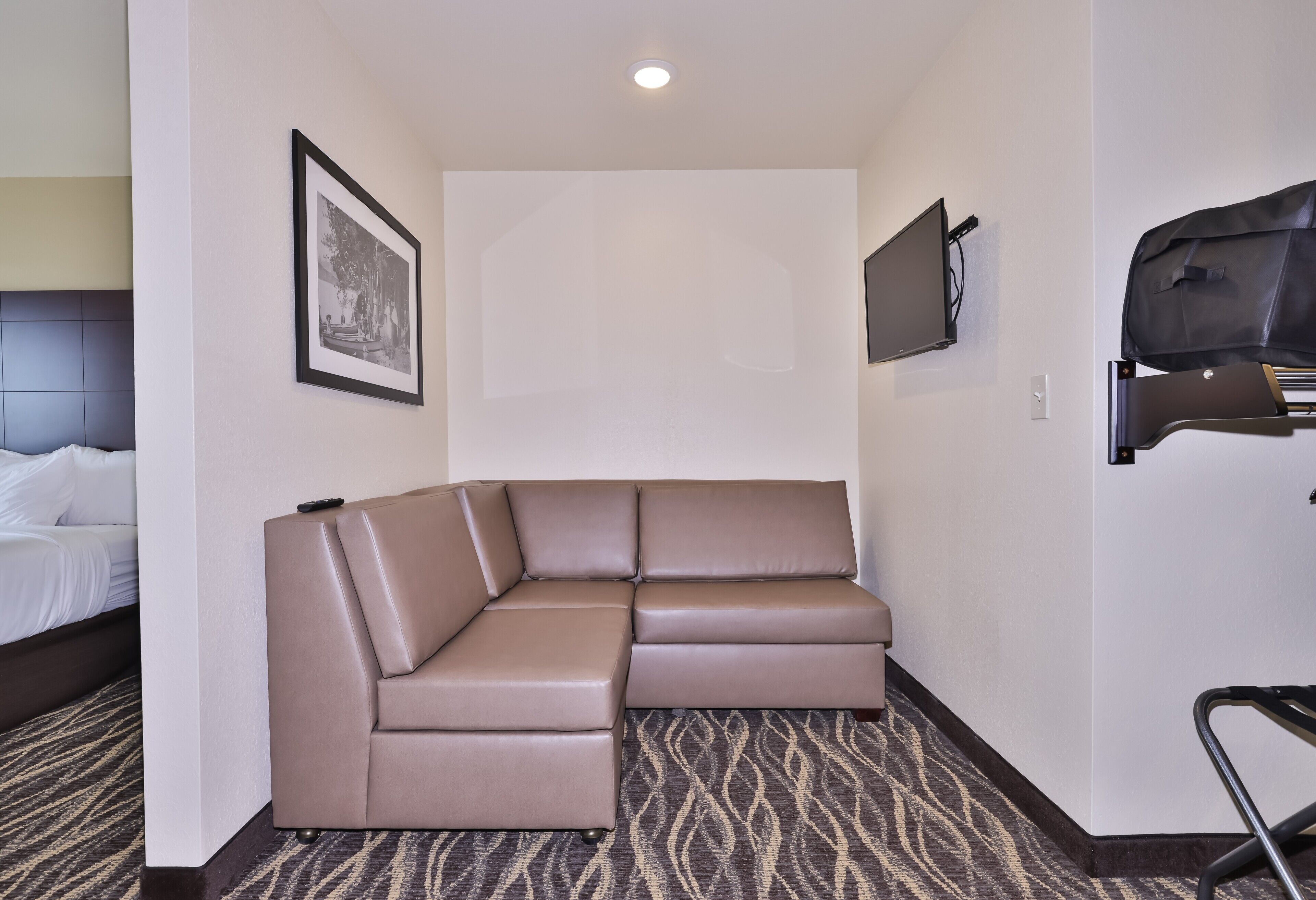 A hotel room with a large L-shaped sectional sofa, a flat-screen TV mounted on the wall, and a bed with white linens.