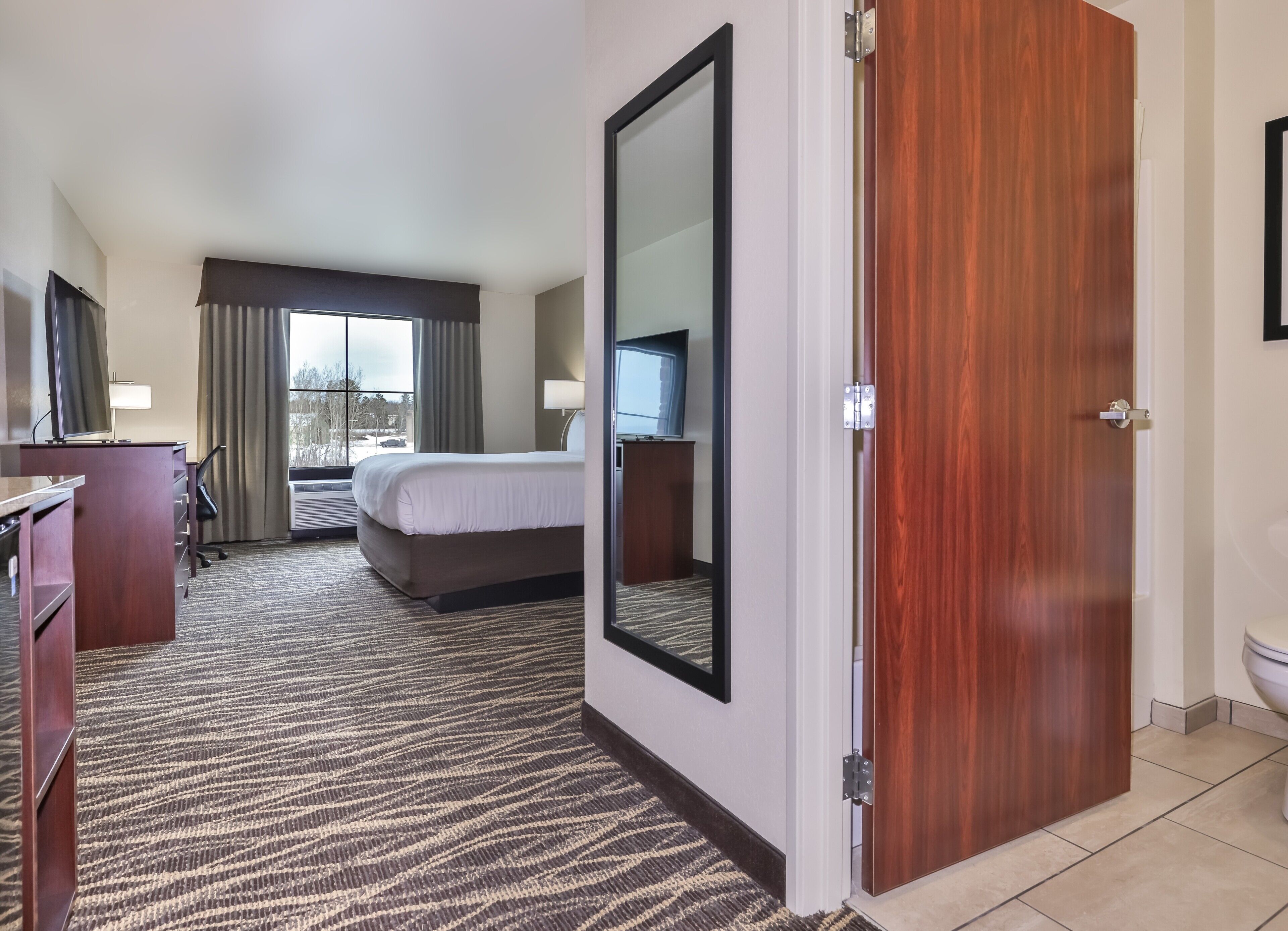 Habitación de hotel con cama, escritorio, silla, televisor y una ventana grande con cortinas.