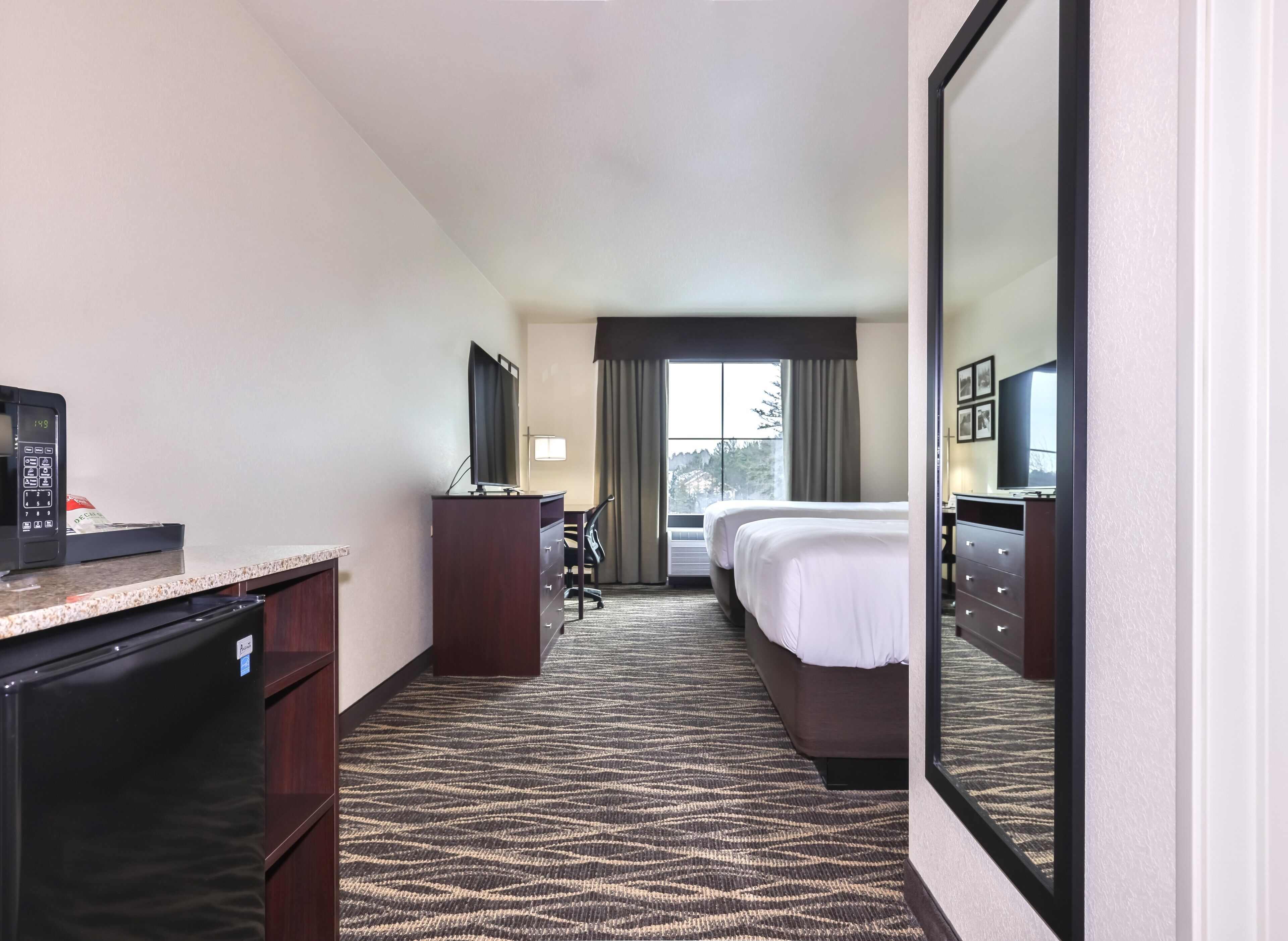 A hotel room with a bed, a desk with a computer, a refrigerator, and a large mirror.