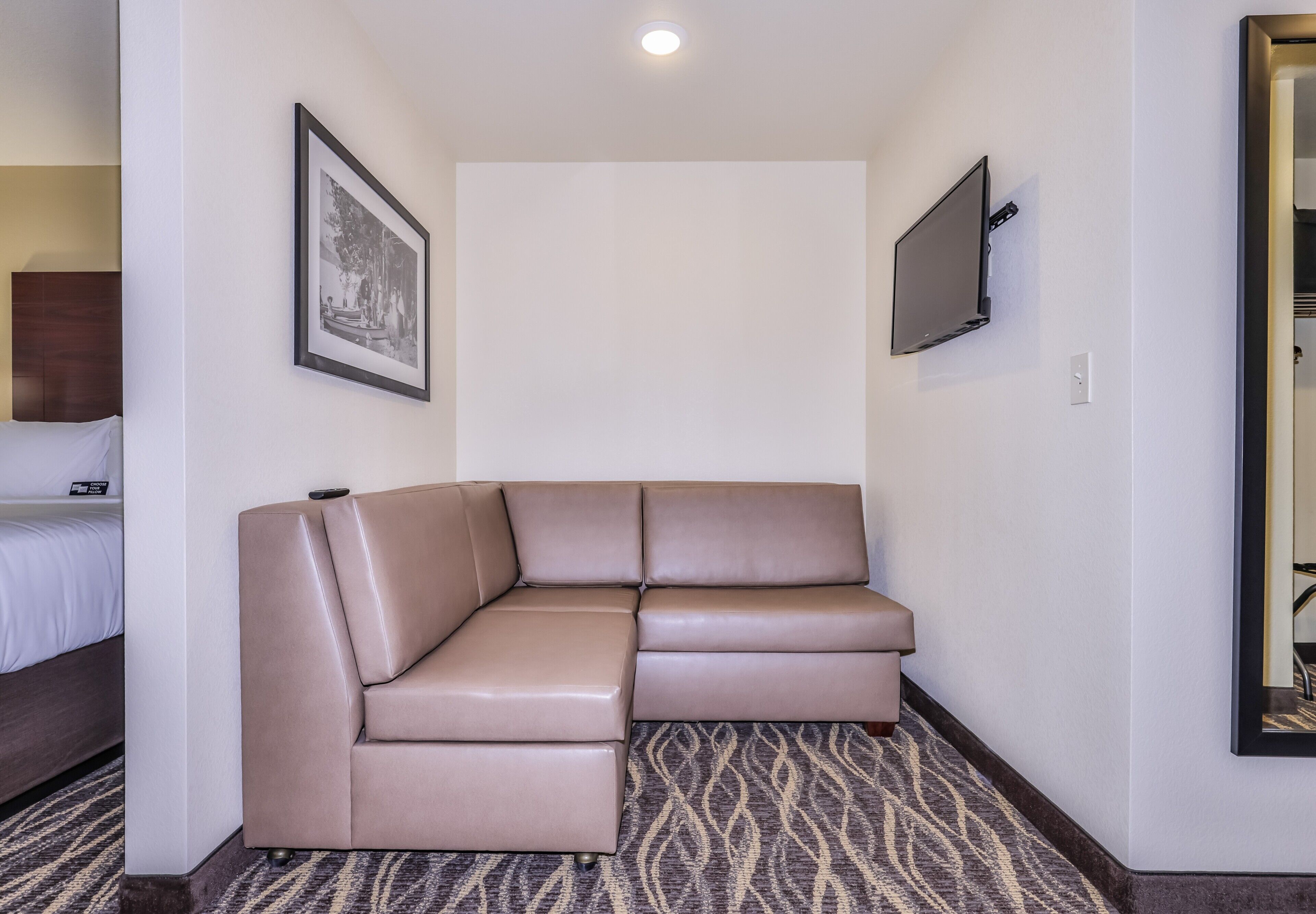 A hotel room with a large L-shaped sectional sofa, a flat-screen TV mounted on the wall, and a patterned carpet.