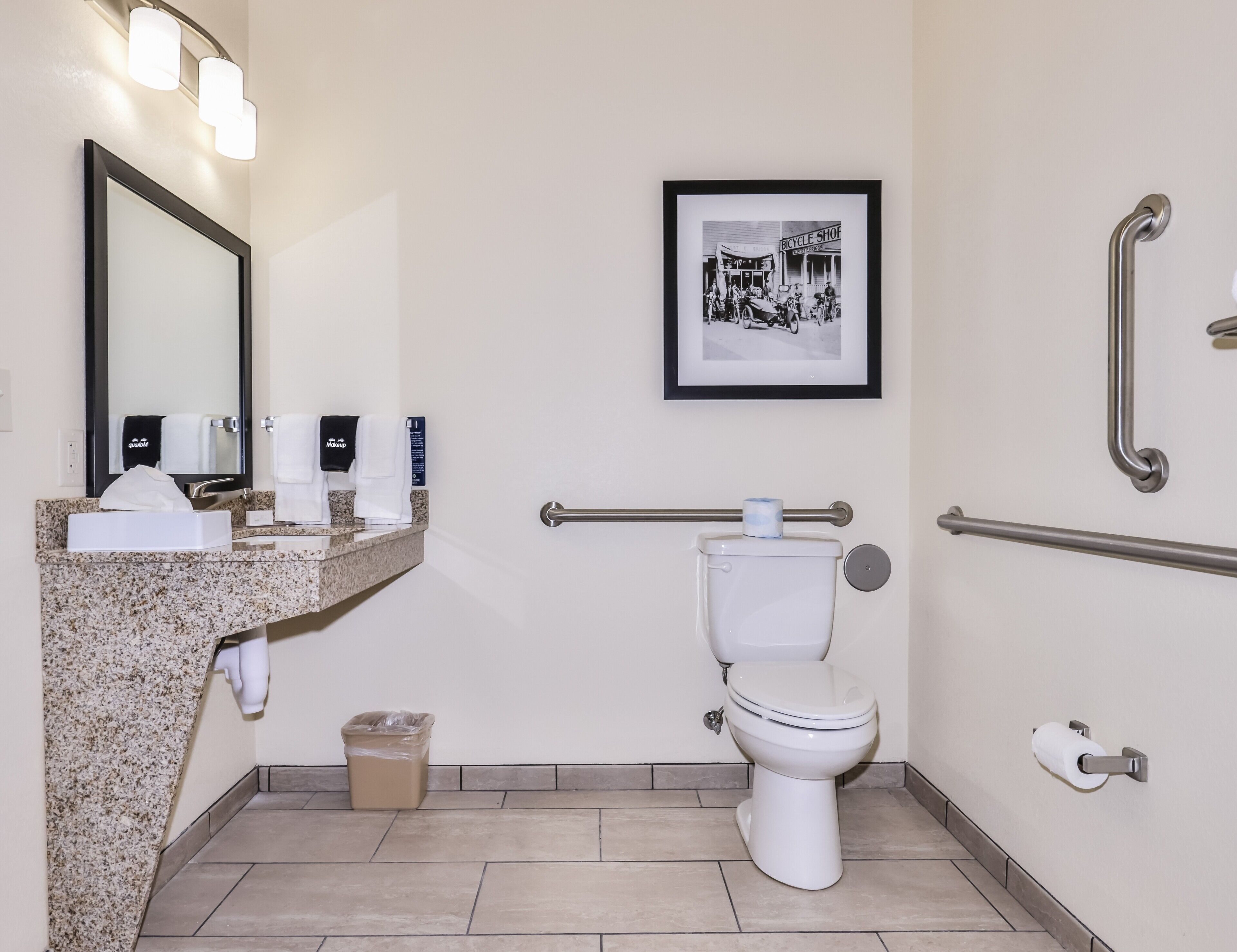 A modern bathroom with a granite countertop, a toilet, a hand dryer, a trash can, and a framed picture on the wall.