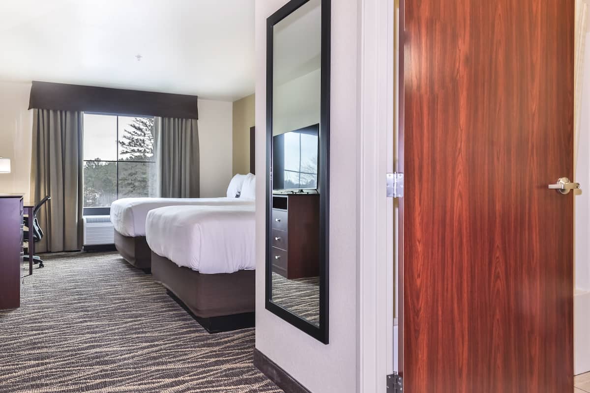 A hotel room with two beds, a large mirror, a desk, and a view of the outdoors.