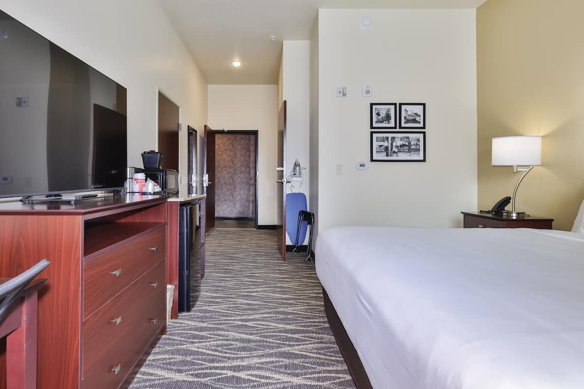 A hotel room with a bed, a desk with a computer, a chair, a lamp, and a door leading to another room.