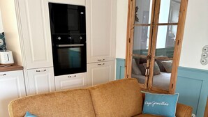 Un sofá marrón tejido con dos almohadones, una cocina con armarios blancos y un horno negro, y una ventana con marco de madera.