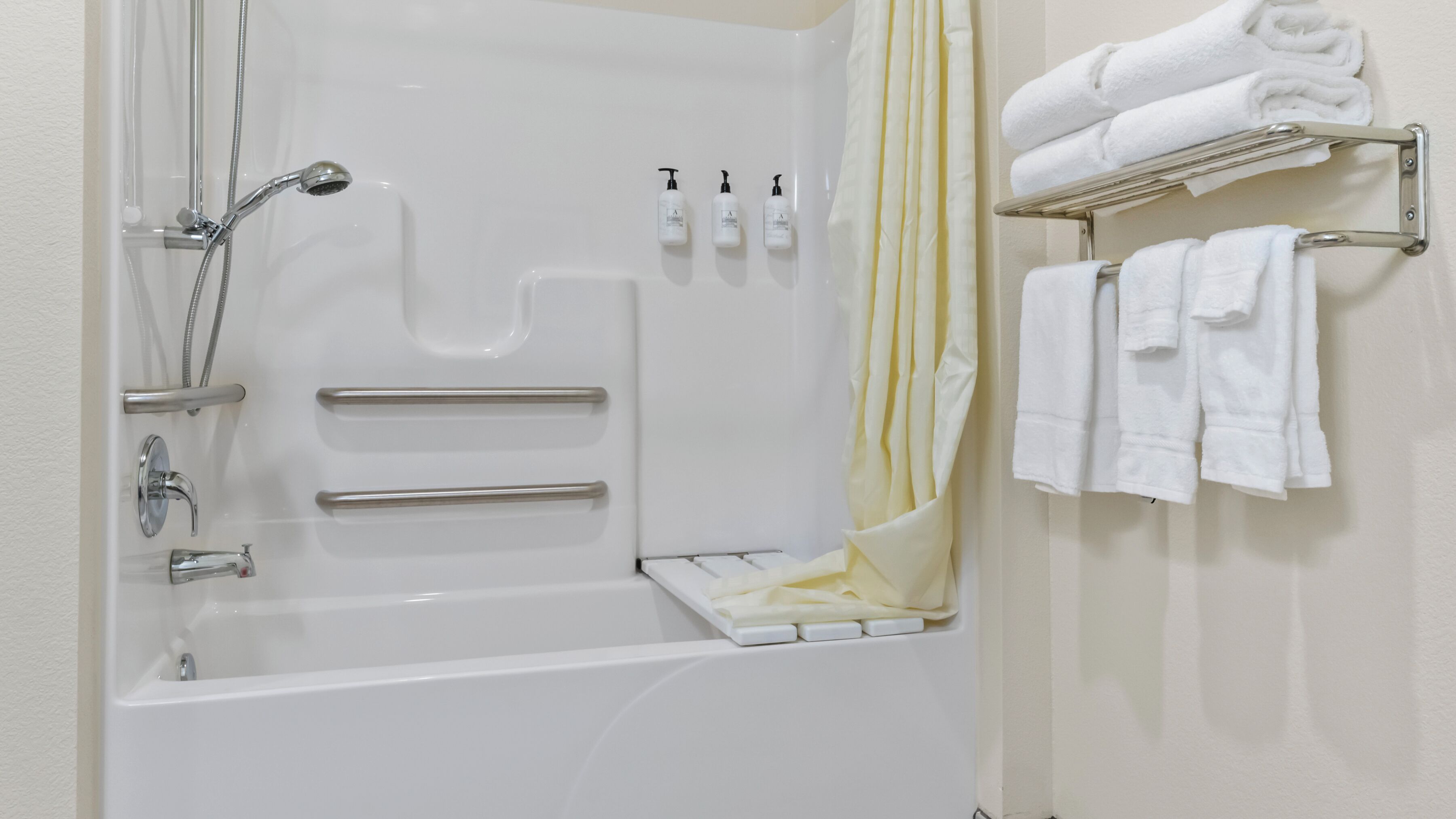 A modern bathroom with a bathtub, shower, and handrails. There are toiletries on the wall and towels on a rack.