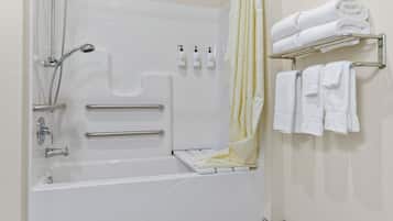 A modern bathroom with a bathtub, shower, and handrails. There are toiletries on the wall and towels on a rack.