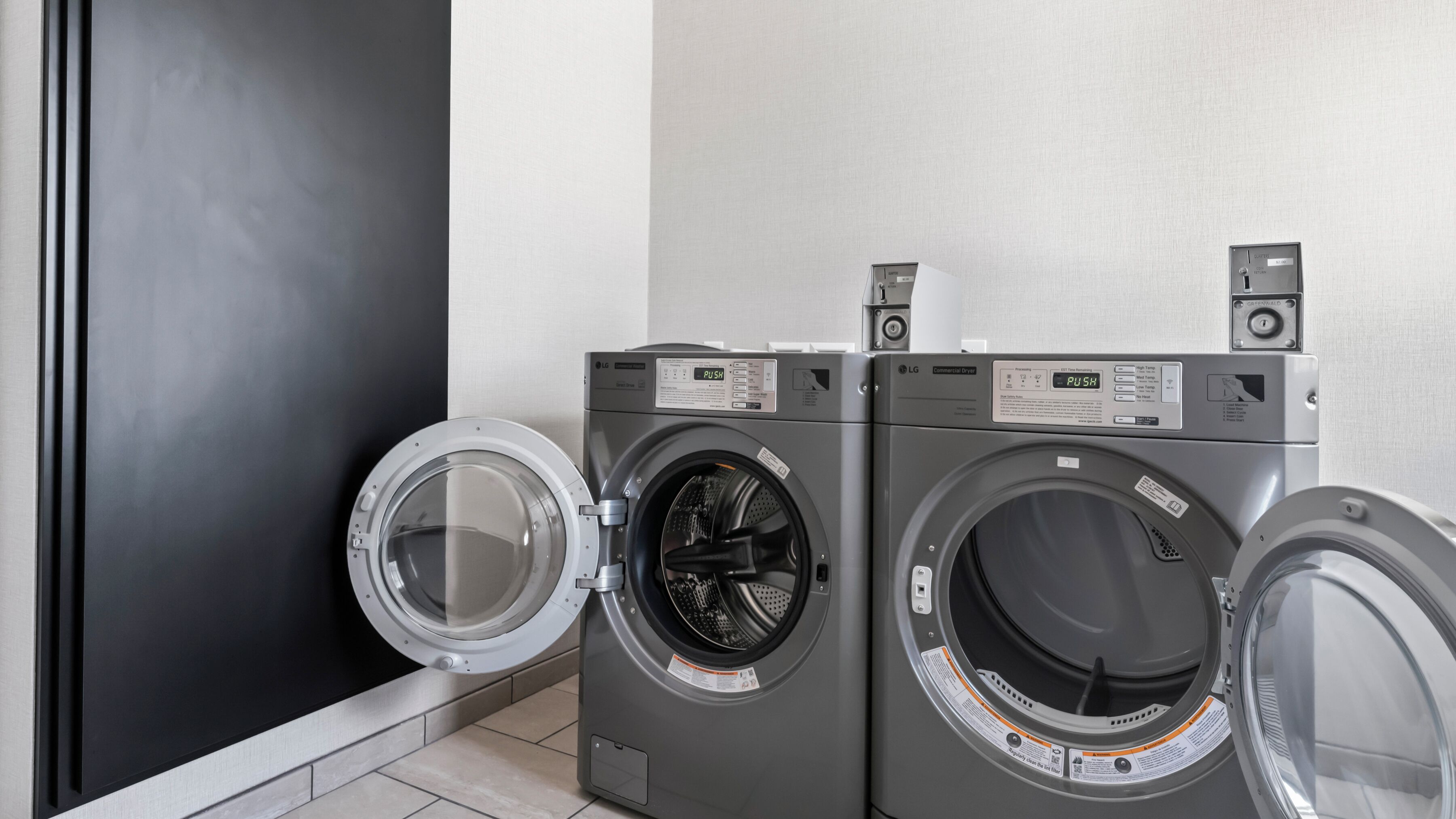 Two front-loading LG commercial dryers in a laundry room with a black door and tiled floor.