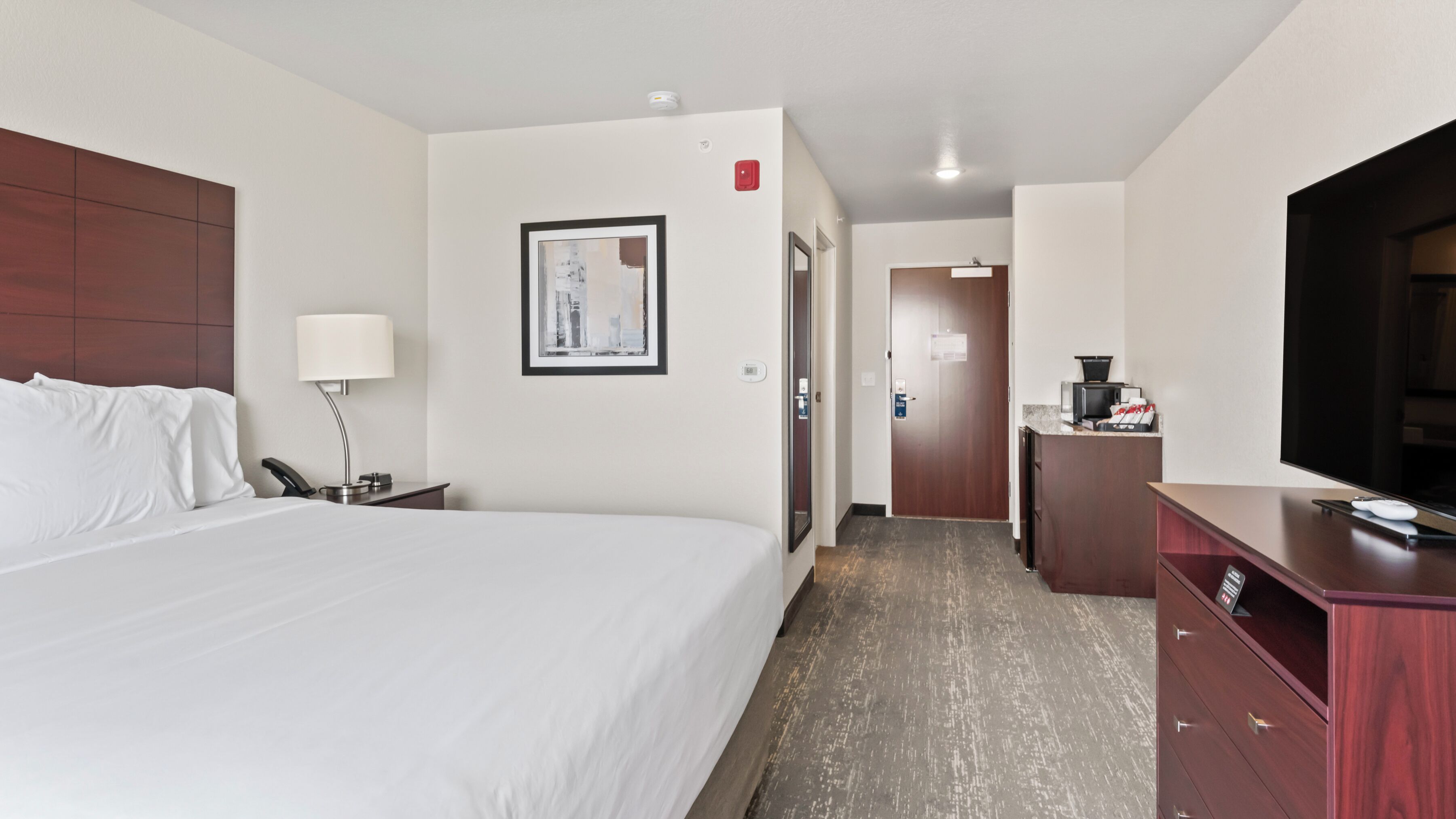 Ein Hotelzimmer mit einem großen Bett, einem Schreibtisch mit Lampe, einem Fernseher und einer kleinen Küchenzeile.