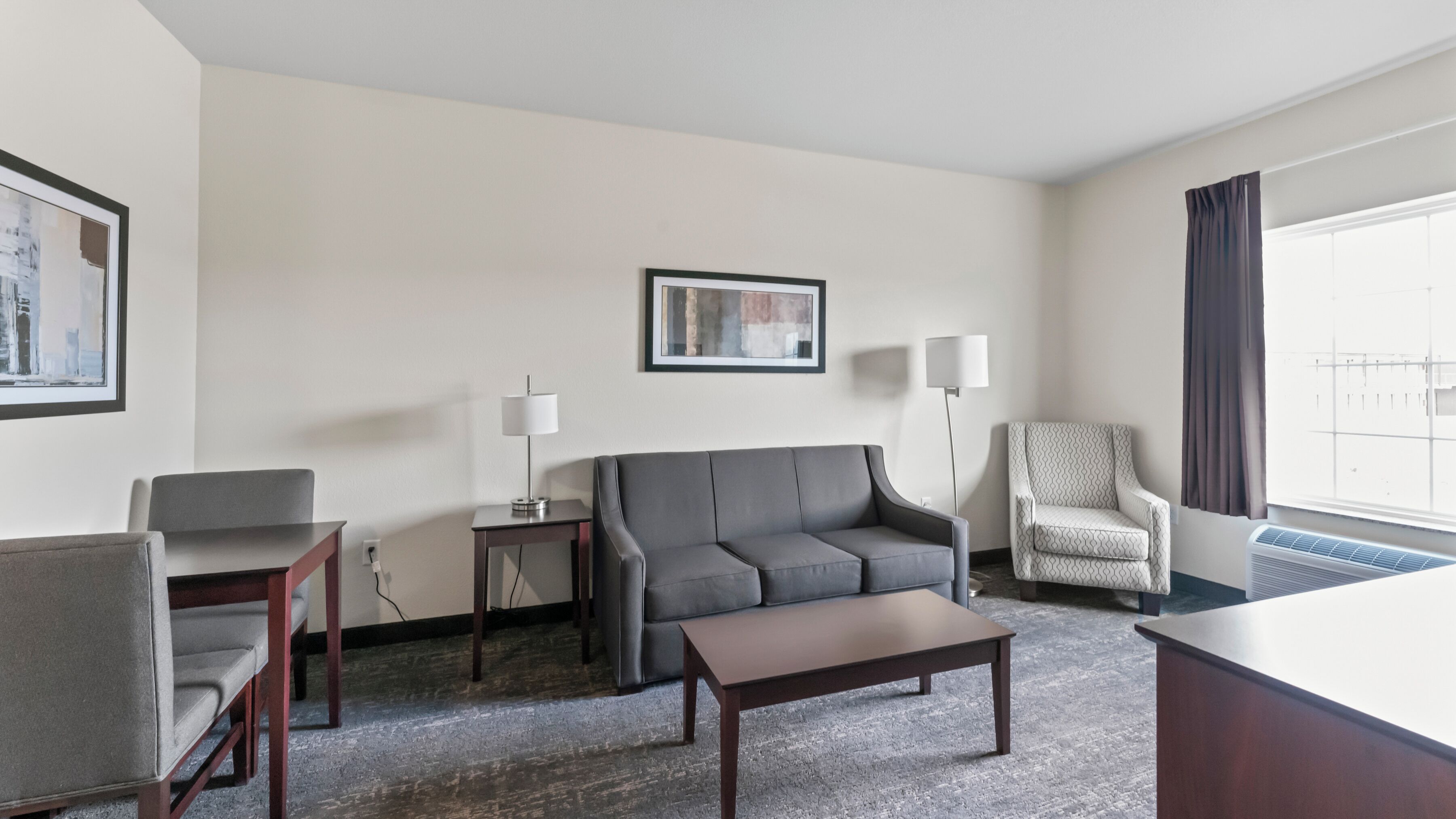A hotel room with a grey sofa, a wooden coffee table, a grey armchair, a patterned armchair, a floor lamp, a side table, and a framed picture on the wall.