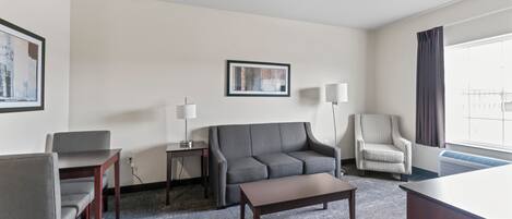 A hotel room with a grey sofa, a wooden coffee table, a grey armchair, a patterned armchair, a floor lamp, a side table, and a framed picture on the wall.