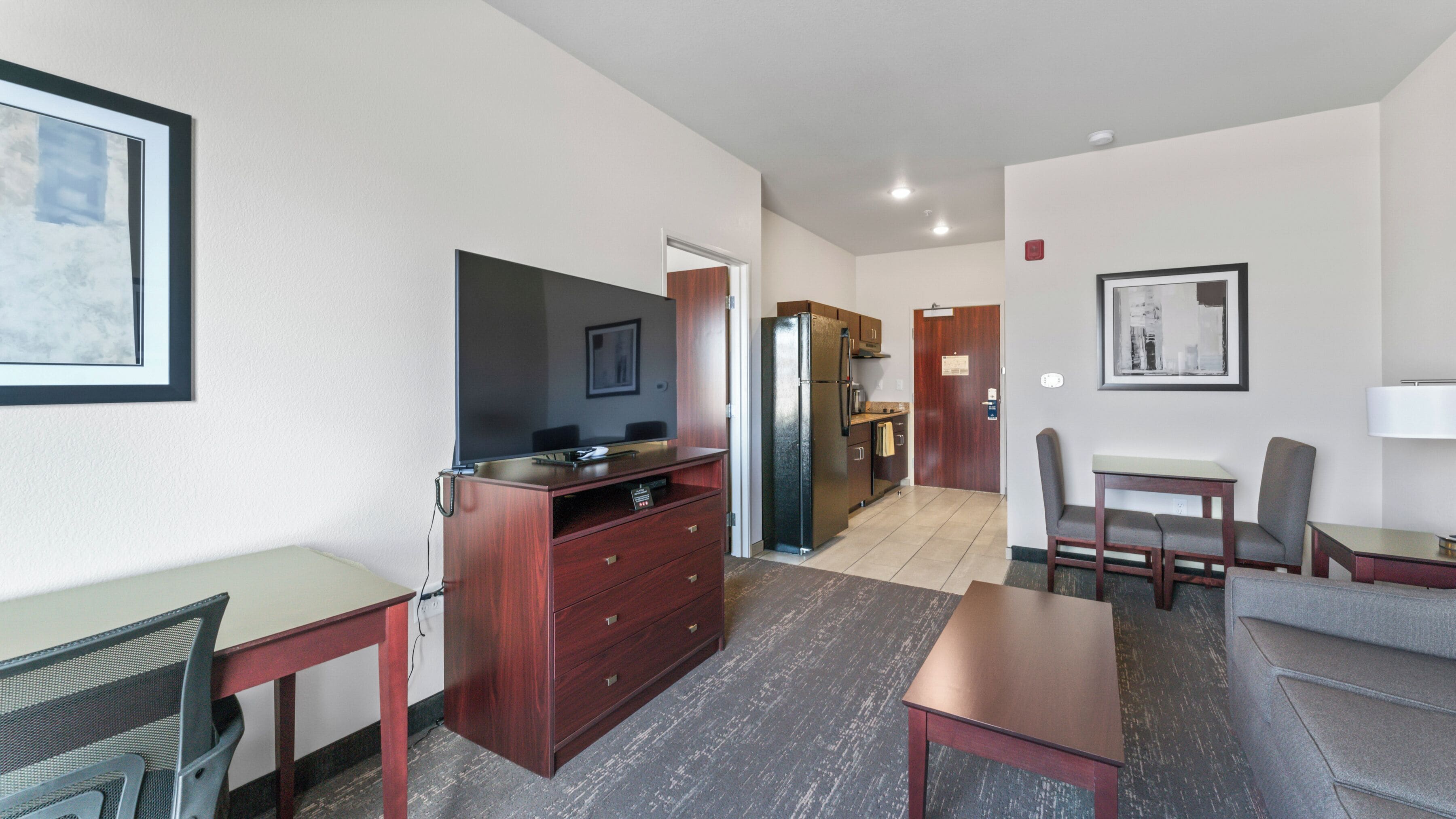 A hotel room with a desk, chair, TV, refrigerator, and a dining area with a table and chairs.