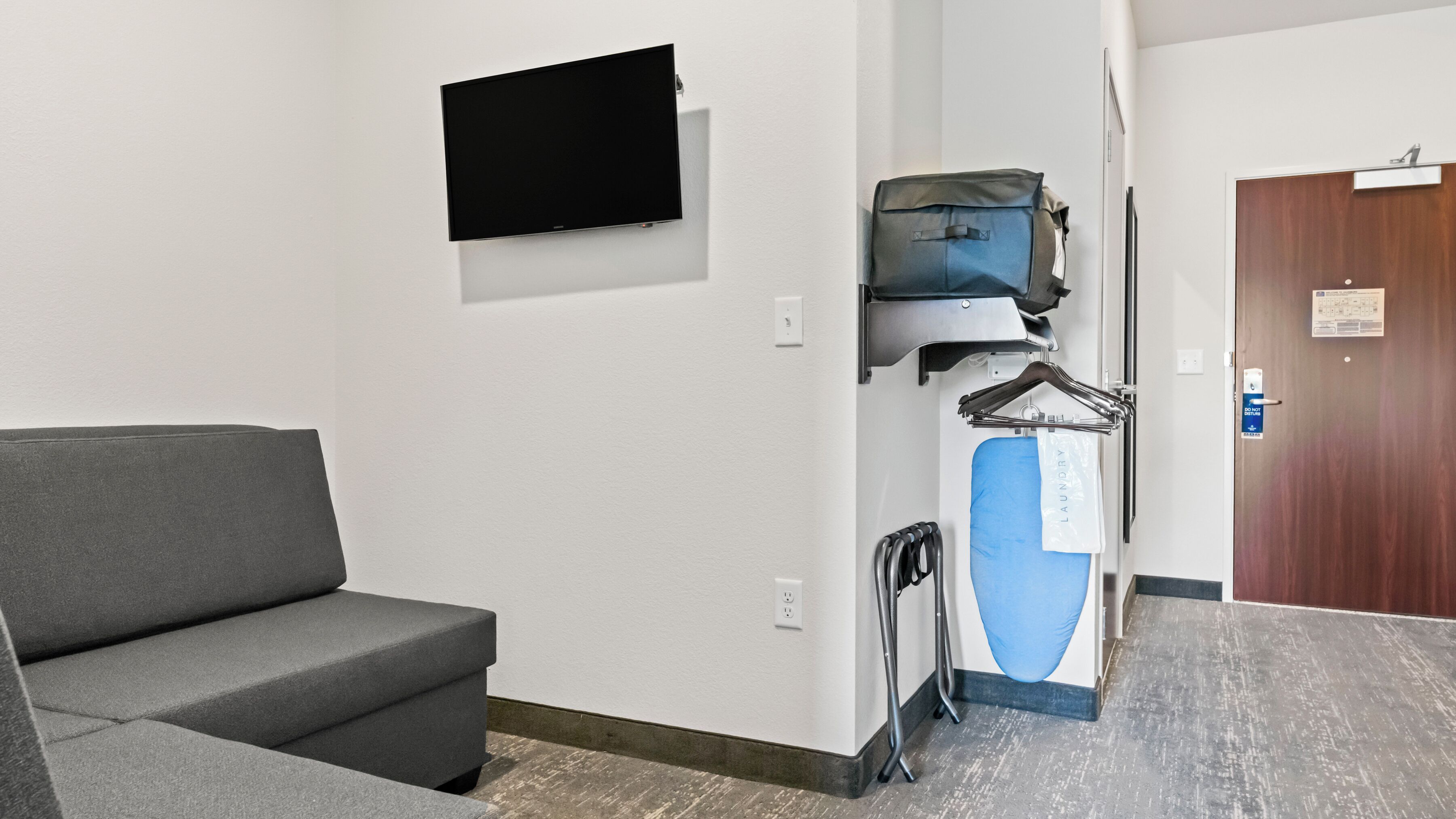 Habitación de hotel con un sofá gris, un televisor de pantalla plana montado en la pared, un cesto azul para ropa y una puerta de madera.