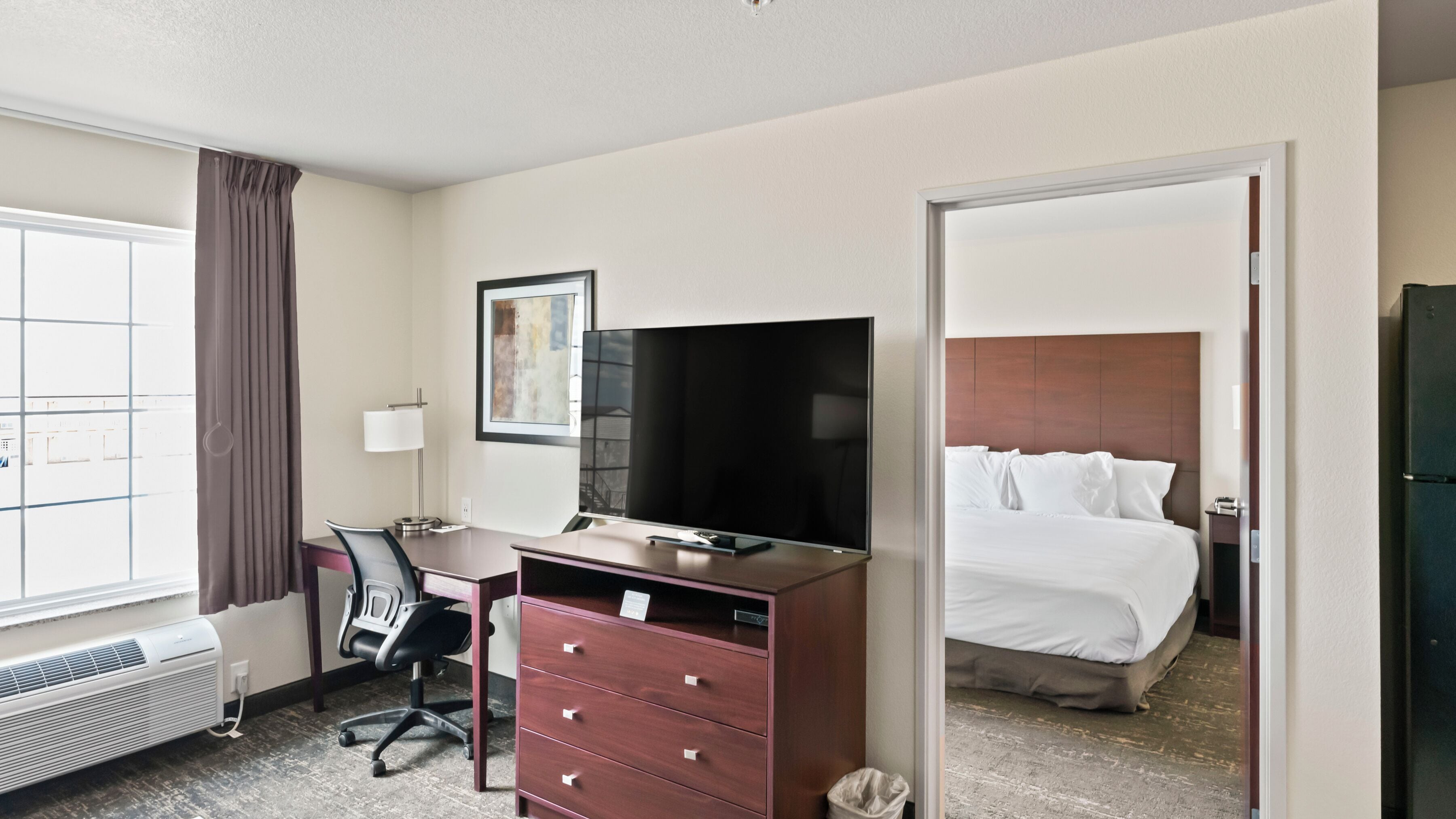 A hotel room with a bed, a desk with a chair, a television, a dresser, and a window with curtains.
