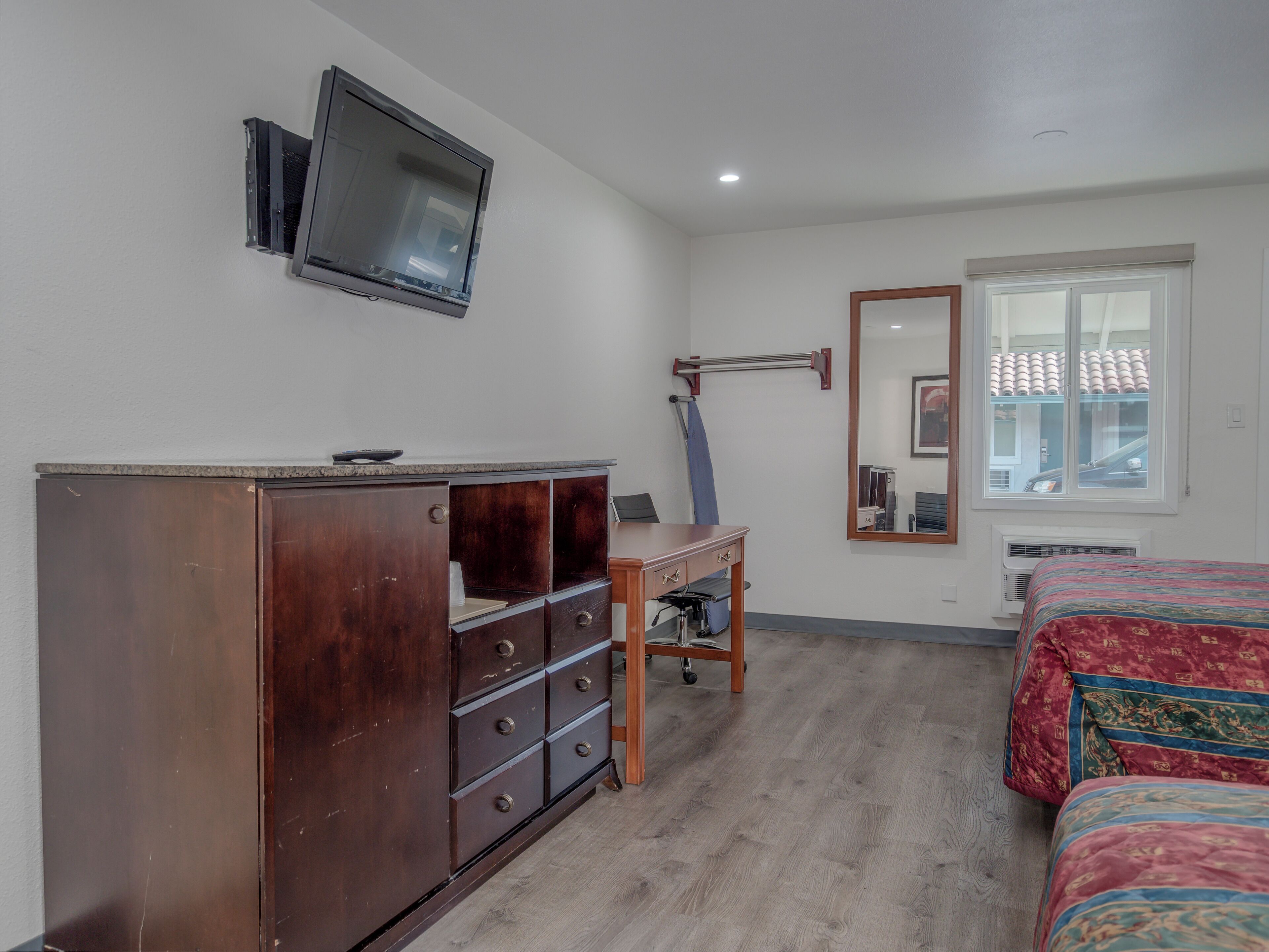 Ein Schlafzimmer mit einem Holzkasten, einem Schreibtisch, einem Stuhl, einem Bett mit gemusterten Textilien und einem an der Wand befestigten Fernseher sowie einem Fenster mit Jalousien.