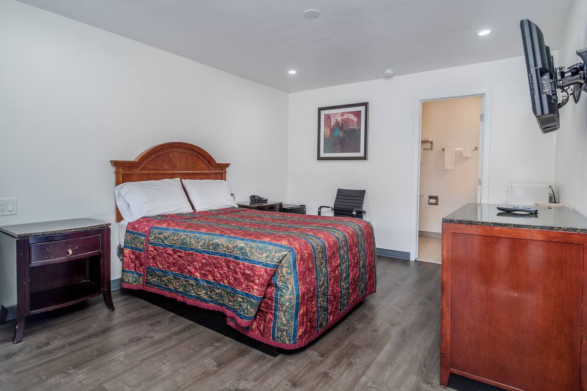 Habitación de hotel con cama de madera, un escritorio, una silla, una lámpara de escritorio y un cuadro en la pared.
