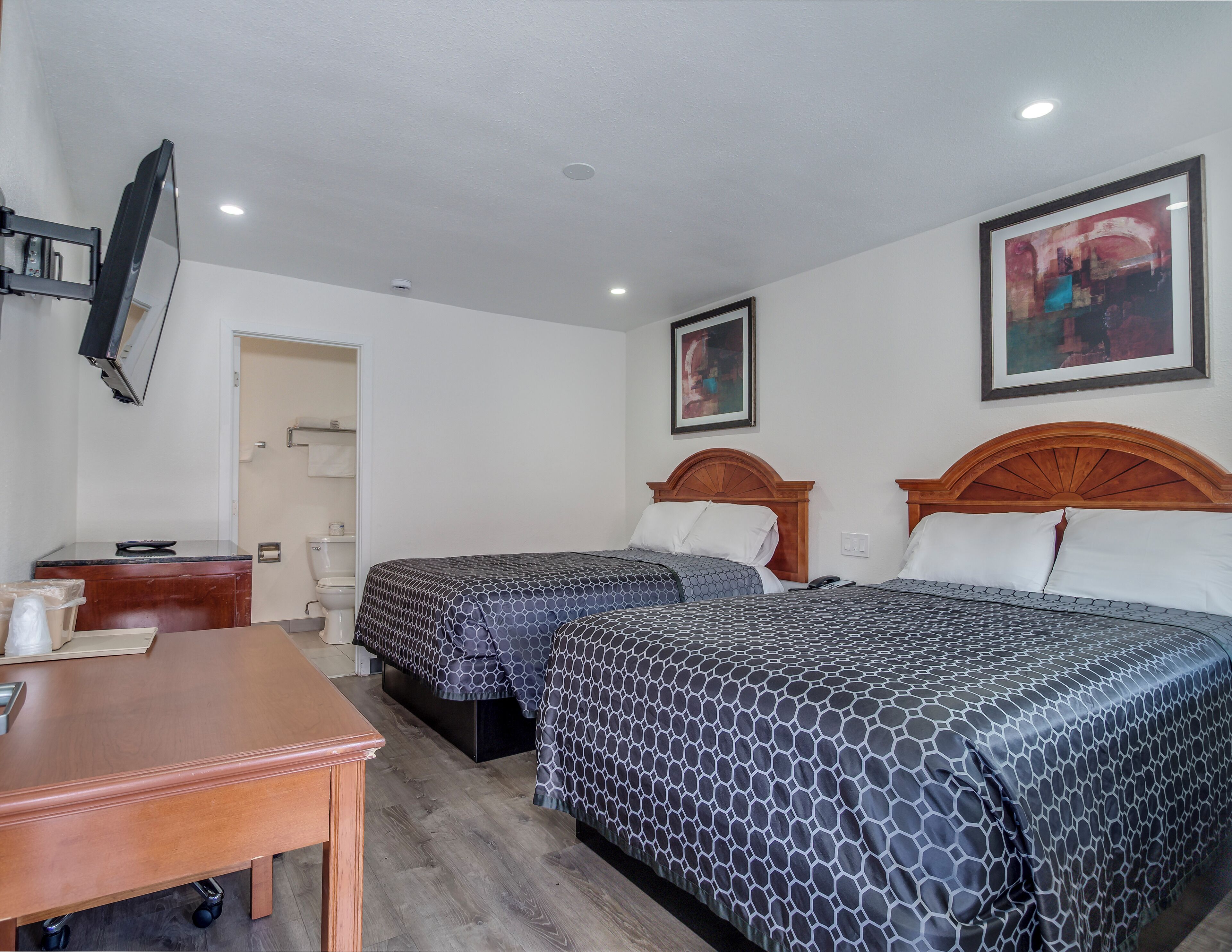 Quarto de hotel com duas camas, uma escrivaninha de madeira, televisão e banheiro visível por uma porta aberta.
