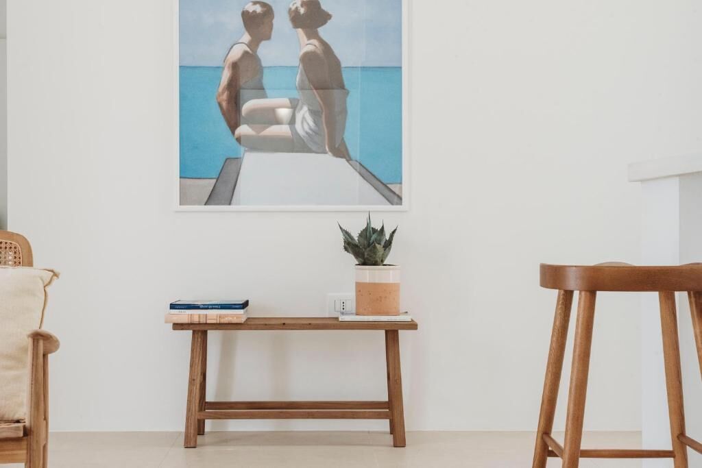 A room with a wooden bench, a small table with a plant and books, and a framed painting of two people by the sea.