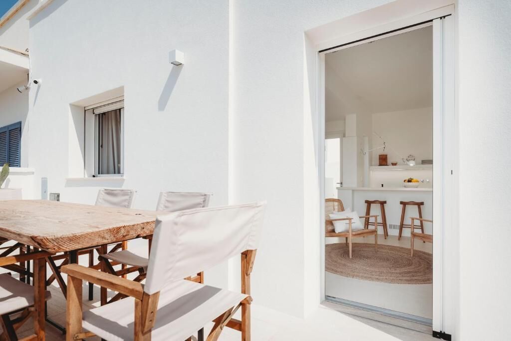 A white villa with a wooden dining table and chairs, a kitchen visible through an open door, and a small balcony with a potted plant.