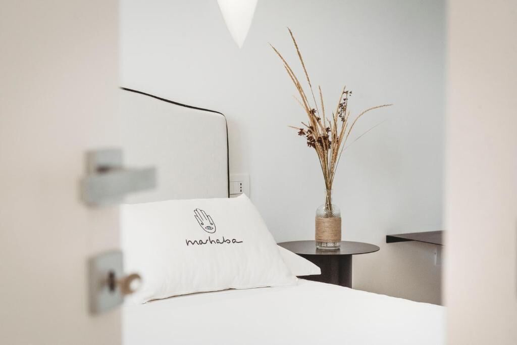 A neatly arranged hotel room with a bed, a bedside table with a vase of dried plants, and a wall-mounted light fixture.