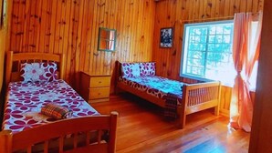 A room with wooden walls and floors, two single beds with floral bedding, a small table, and a window with curtains. - ROOMS AT AN AMERICAN-STYLE CABIN (Sagada)
