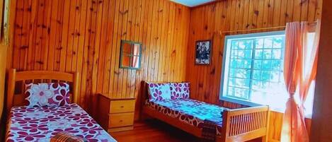 A room with wooden walls and floors, two single beds with floral bedding, a small table, and a window with curtains.