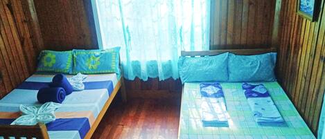 Dos camas individuales con ropa de cama azul y verde, cabeceros de madera y una ventana con cortina blanca.