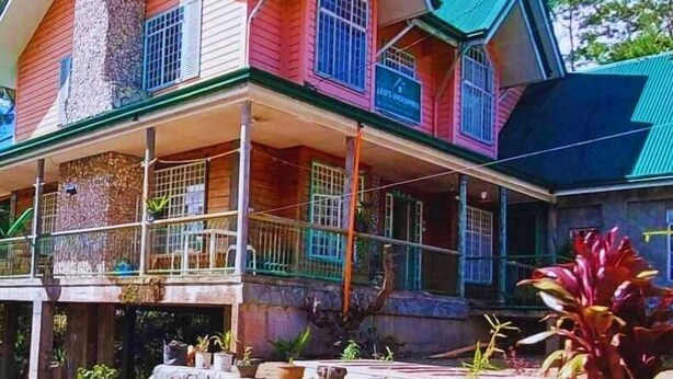 A two-story house with a stone chimney, a covered porch with railings, and a sign indicating a 'Guest House'. - ROOMS AT AN AMERICAN-STYLE CABIN (Sagada)