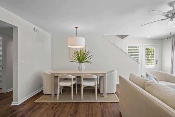 A modern living room with a dining table, chairs, a sofa, a hanging light fixture, and a ceiling fan.