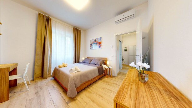 A hotel room with a bed, a desk, a chair, a wooden table, a vase with flowers, and a painting on the wall. - Four stops from the center of Milan, you will have all the comforts of home! (Milano)