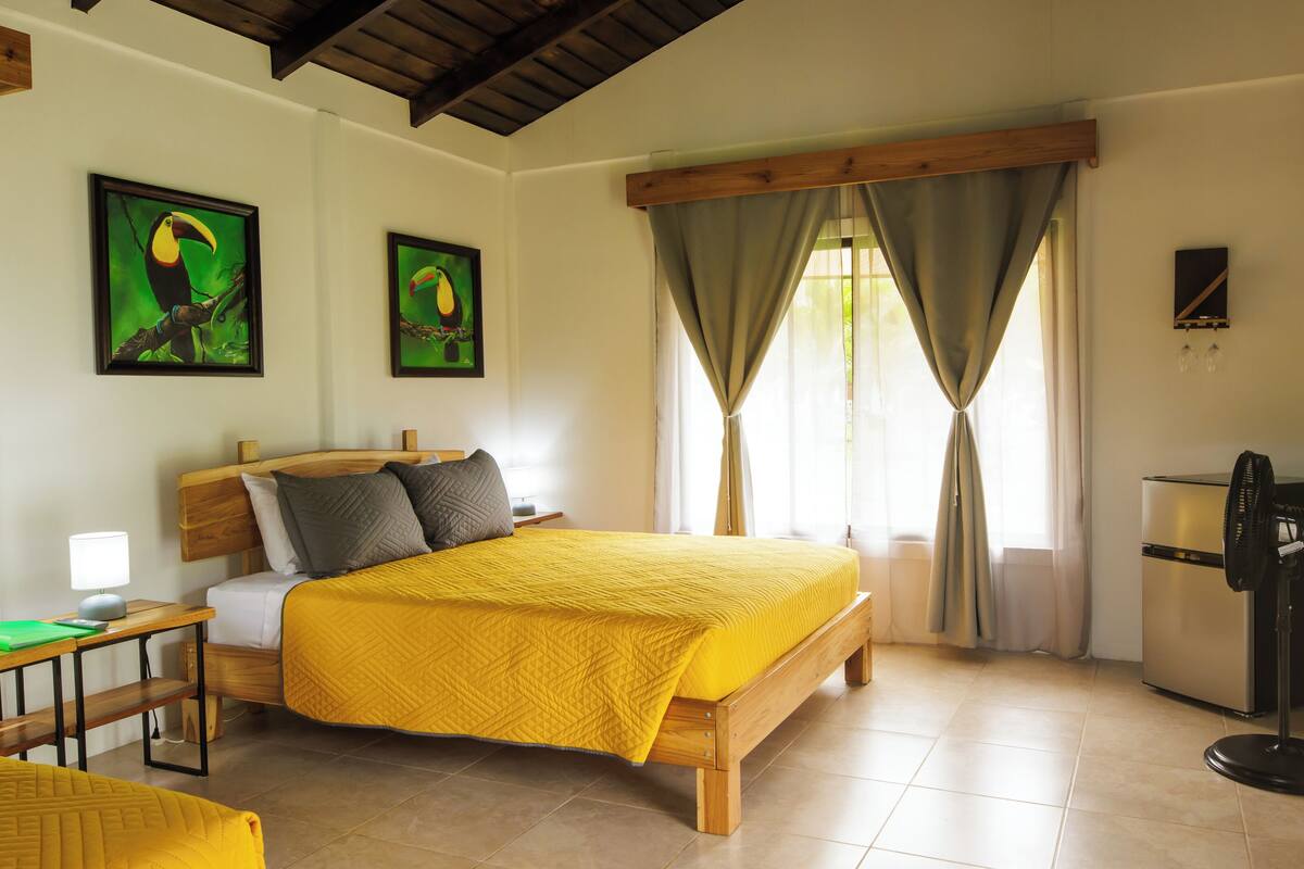 A bedroom with a wooden bed, two paintings of toucans, a desk with a lamp, and a standing fan.
