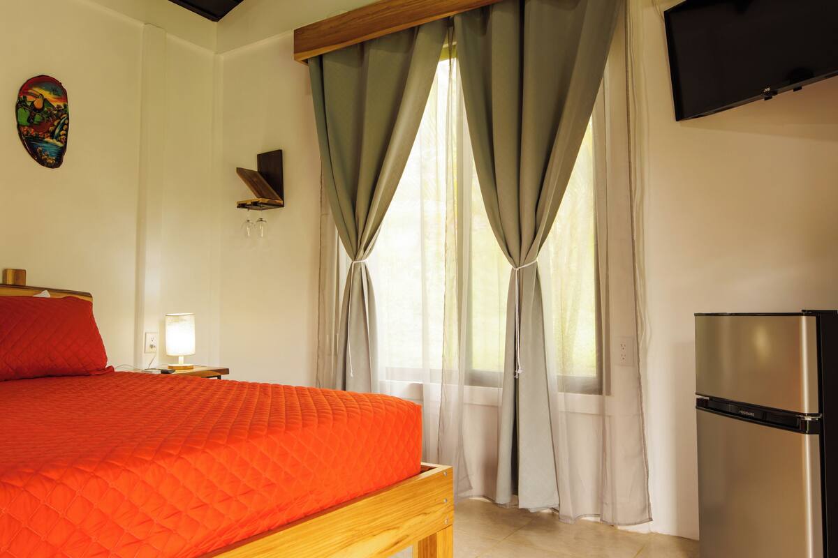 A bedroom with a bed, a nightstand, a lamp, a television, and a refrigerator.