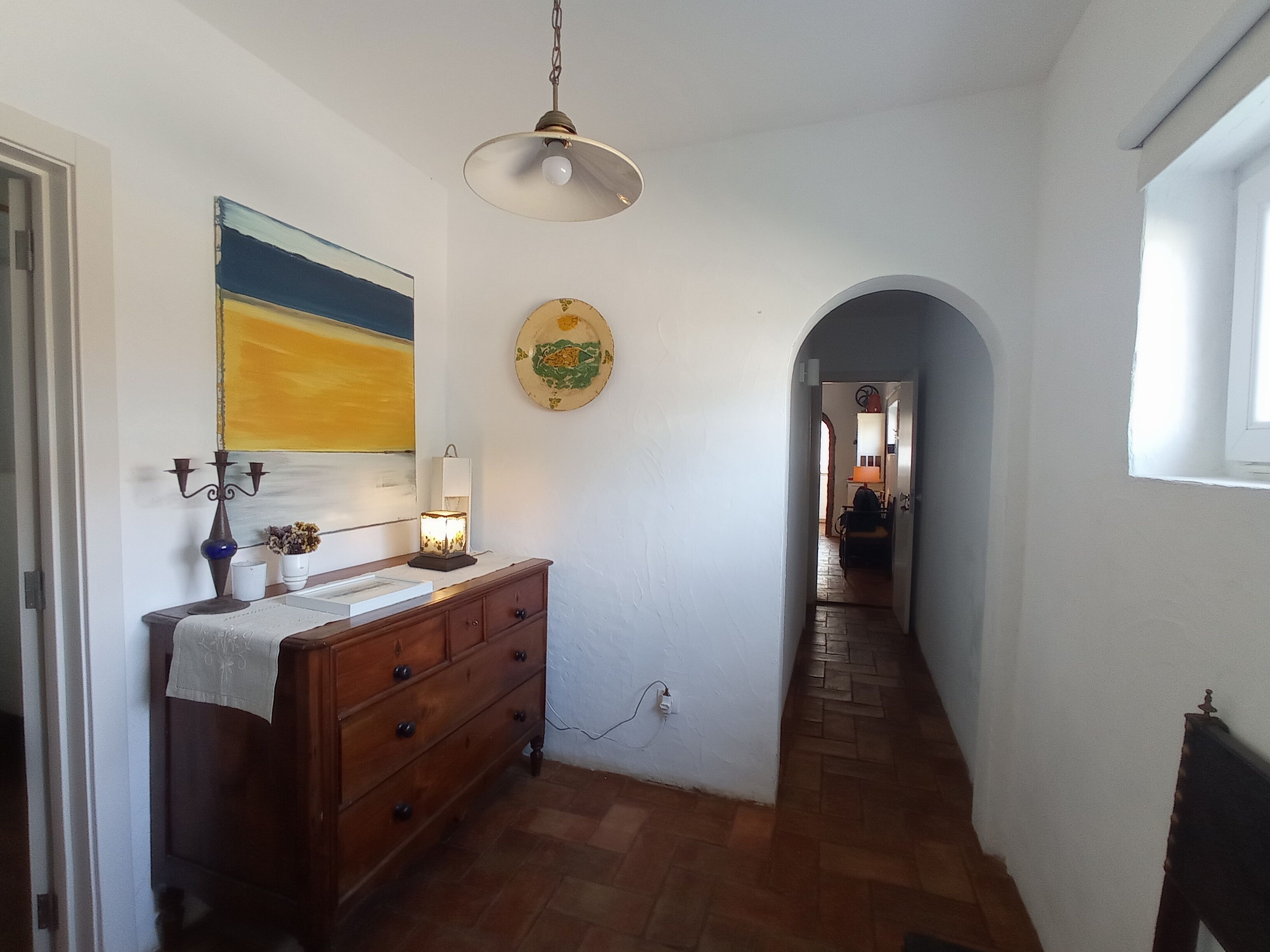 Een gang met een houten dressoir, een hanglamp, een schilderij en een decoratief bord aan de muur.