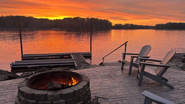 Una terraza junto al lago con sillas Adirondack, un hogar para fuego y un muelle que se adentra en el agua.