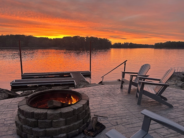 A lakeside deck with Adirondack chairs, a fire pit, and a dock extending into the water.