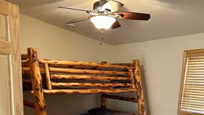 Una cama litera con estructura de madera, un ventilador de techo y una ventana con persianas.